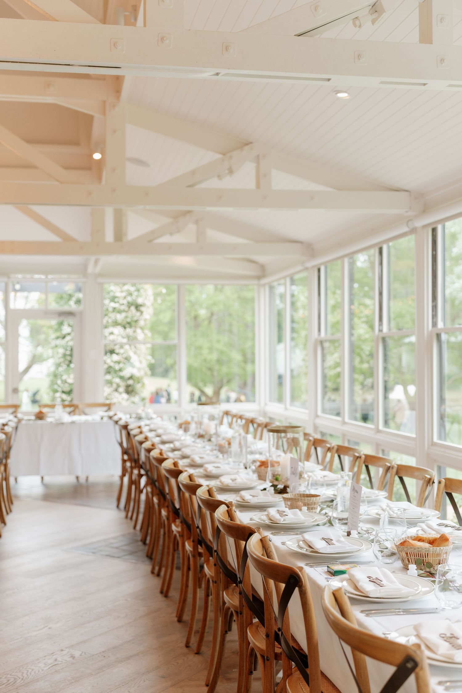Elegant wedding reception hall with long dining tables, wooden cross-back chairs, white linens, and floor-to-ceiling windows.