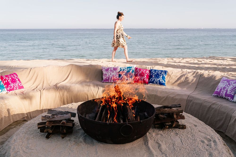 Woman walking on beach near a fire pit with colorful pillows on sandy lounge seating by the ocean.