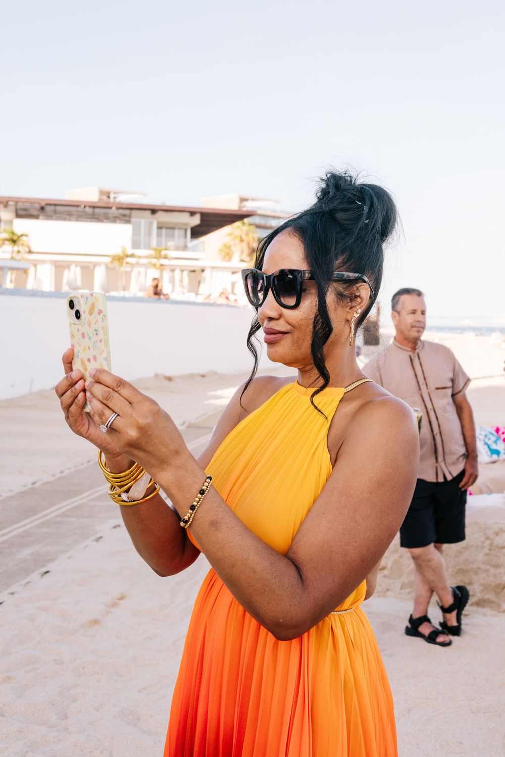 Woman in orange dress taking selfie on beach, wearing sunglasses and gold bracelets, man standing in background.