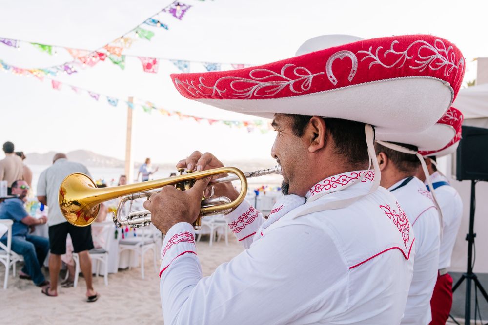 Mariachi musician in traditional sombrero playing trumpet at a festive outdoor beach celebration.