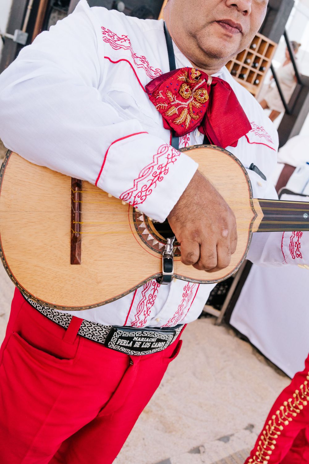 Mariachi musician in traditional white and red embroidered charro outfit playing a vihuela guitar at an outdoor event.