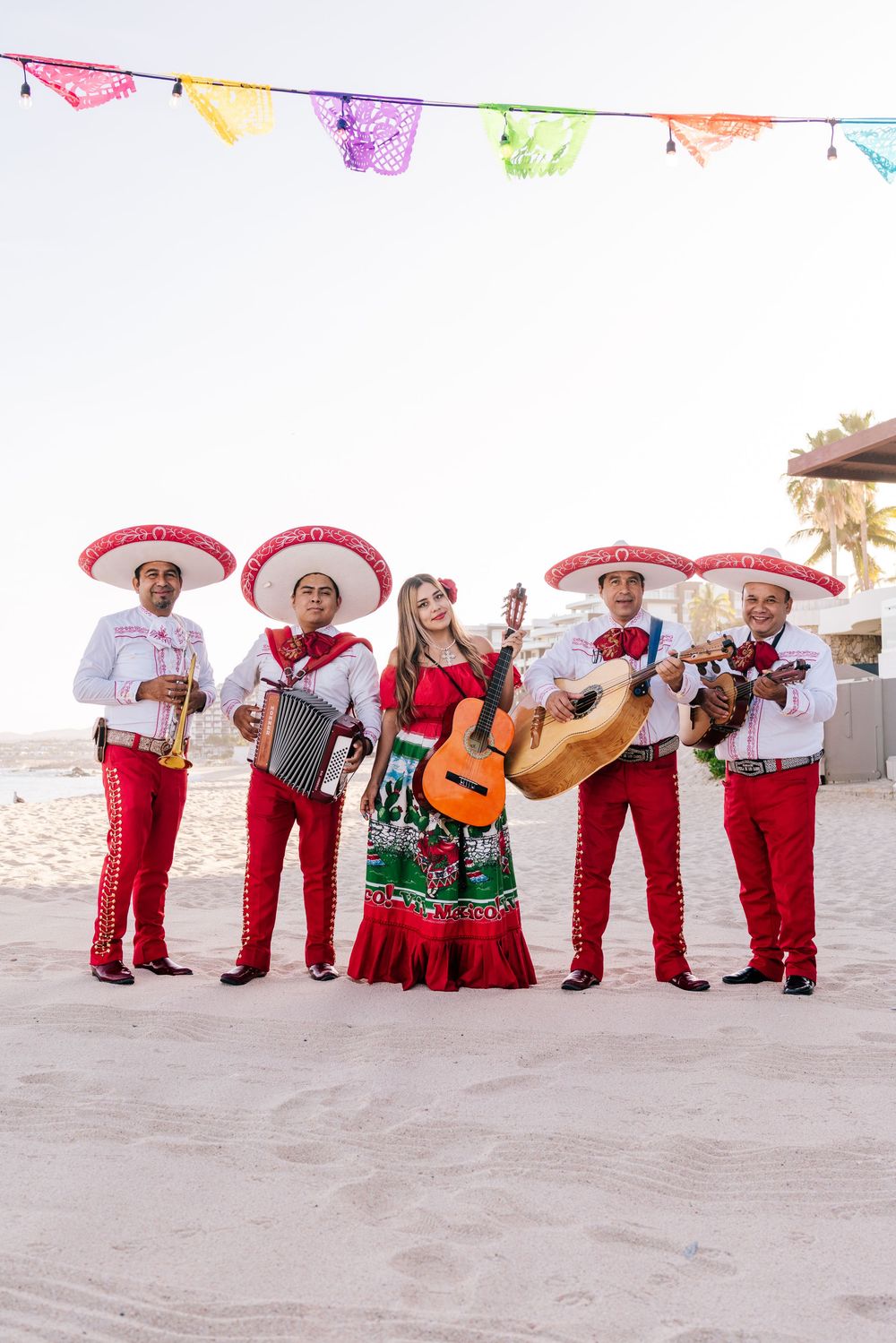 Mariachi band in red charro suits and sombreros performing with a woman in traditional dress on a sunny beach.