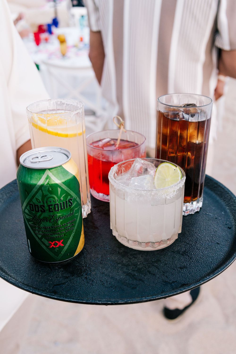 A tray of colorful cocktails including a margarita, cola, red drink, lemonade, and Dos Equis beer can.