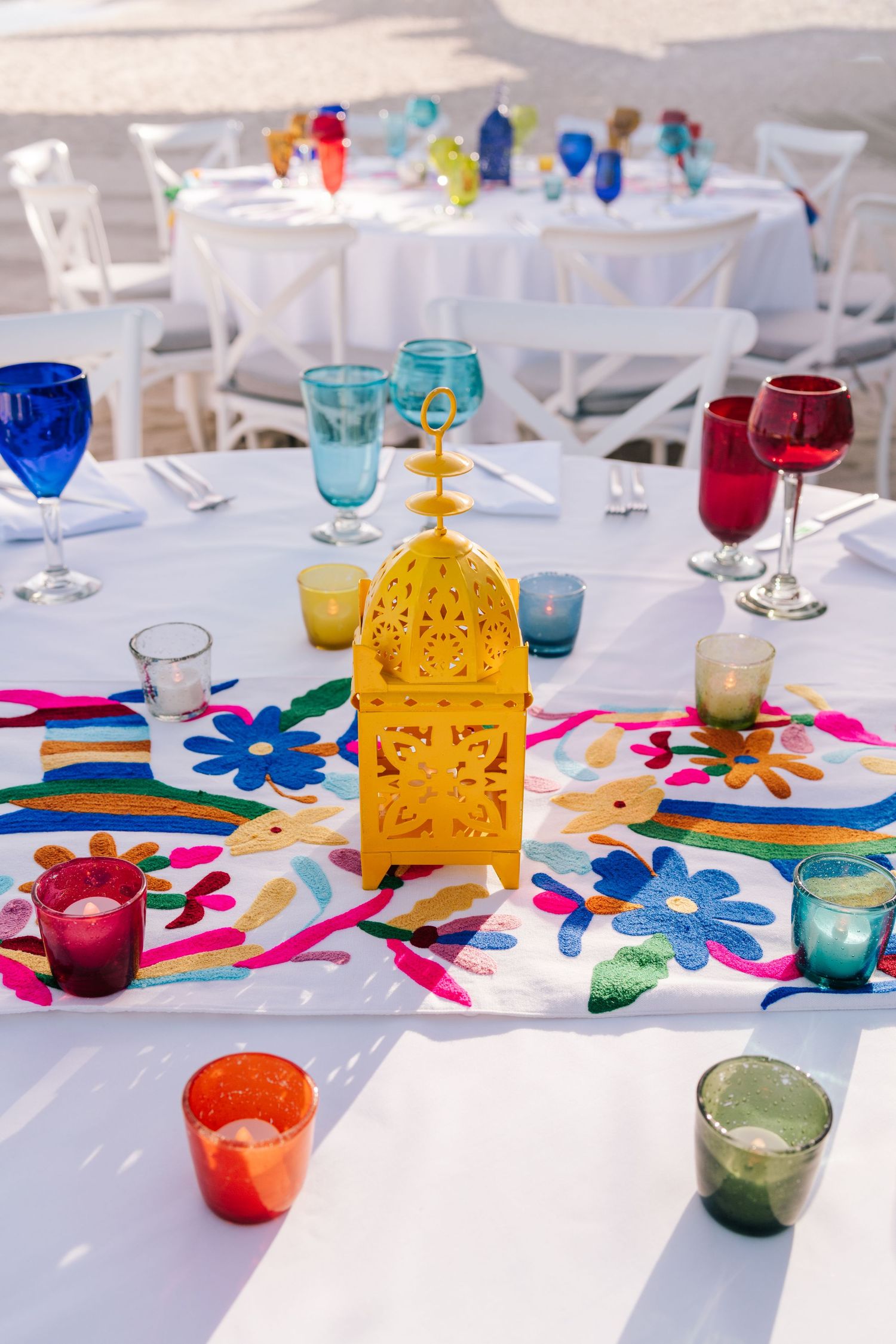Colorful outdoor wedding table with yellow Moroccan lantern, vibrant glassware, floral embroidered runner, and small votive candles.