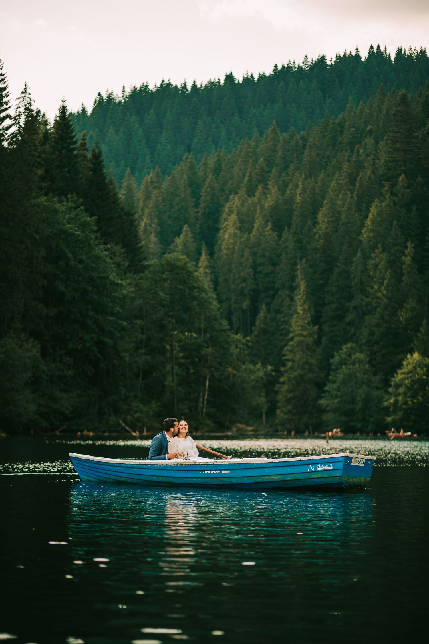 Couple rowing a blue boat on a serene lake surrounded by lush evergreen forest.