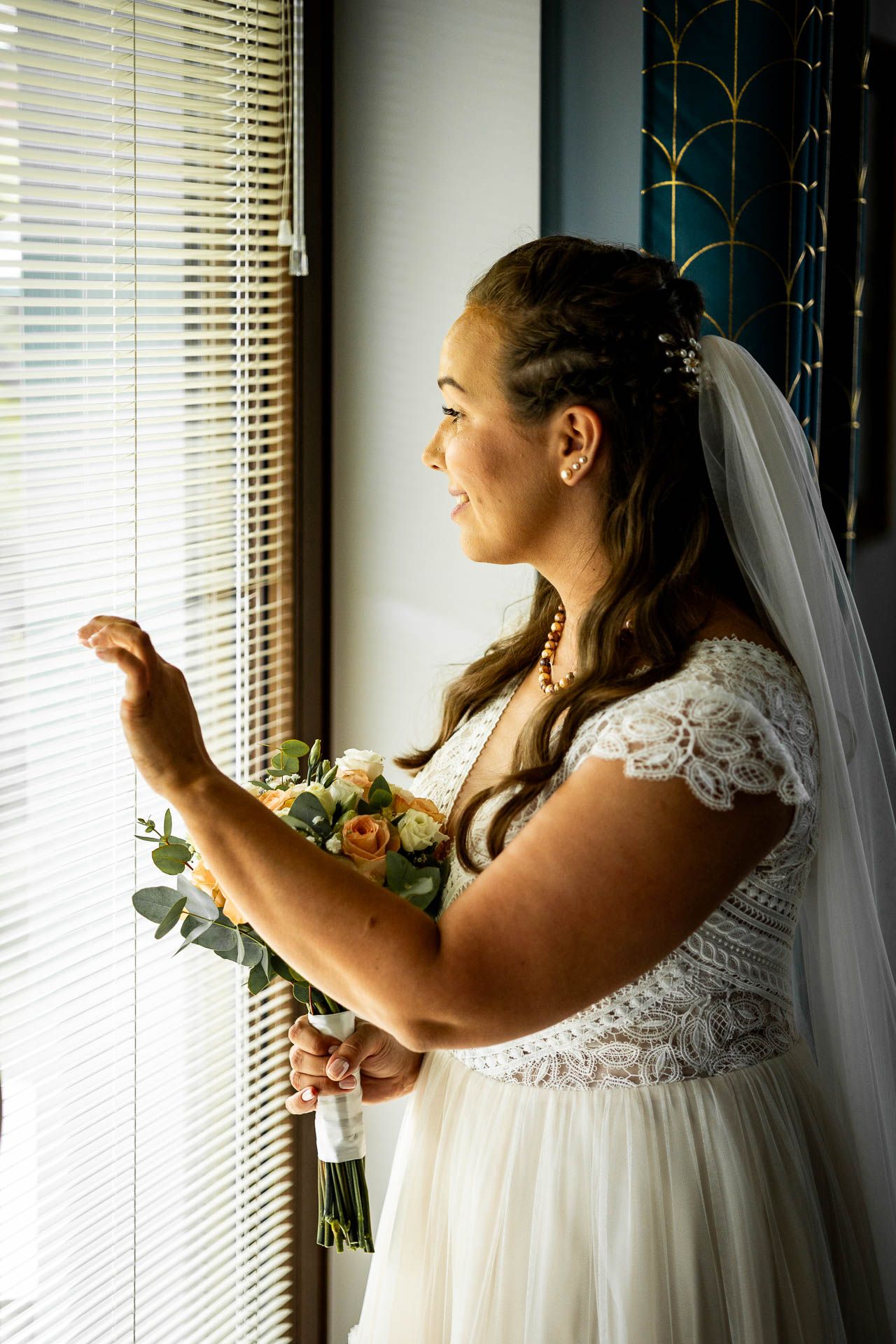Bride in white lace dress looks out window while holding bouquet.