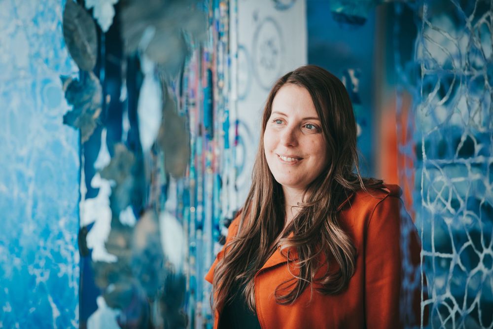 Person in bright orange coat poses against textured blue wall with artistic graffiti elements in urban setting.