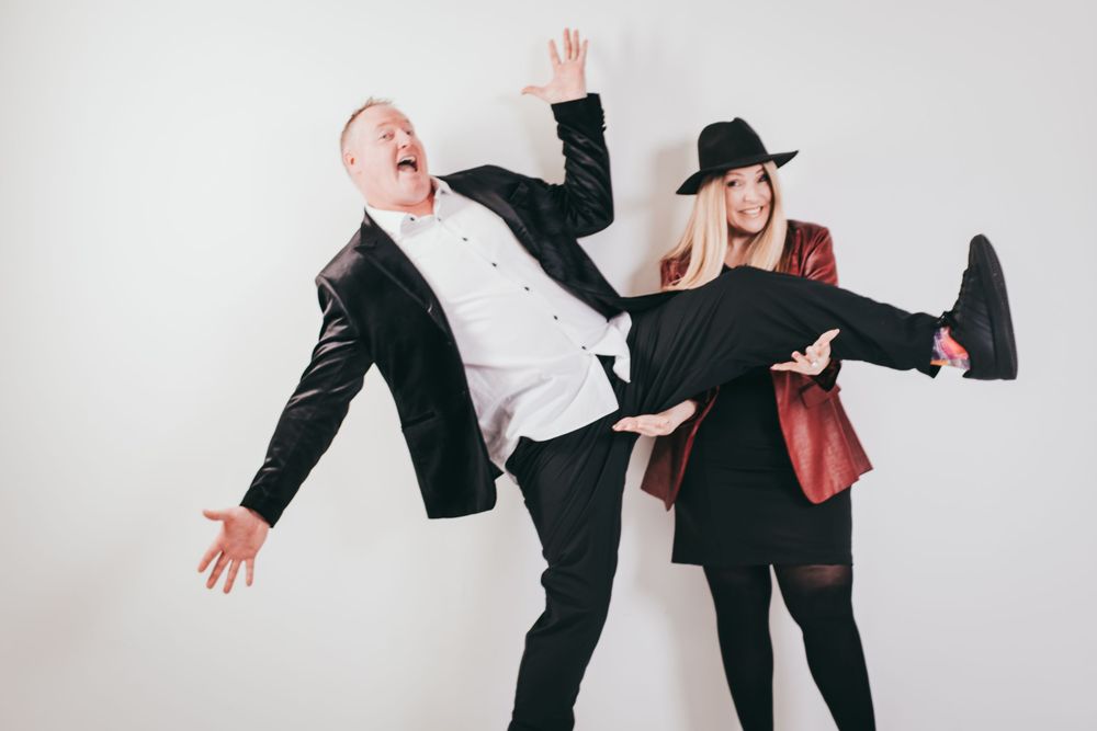 Two people in business attire strike playful poses against a white background for a creative studio portrait.
