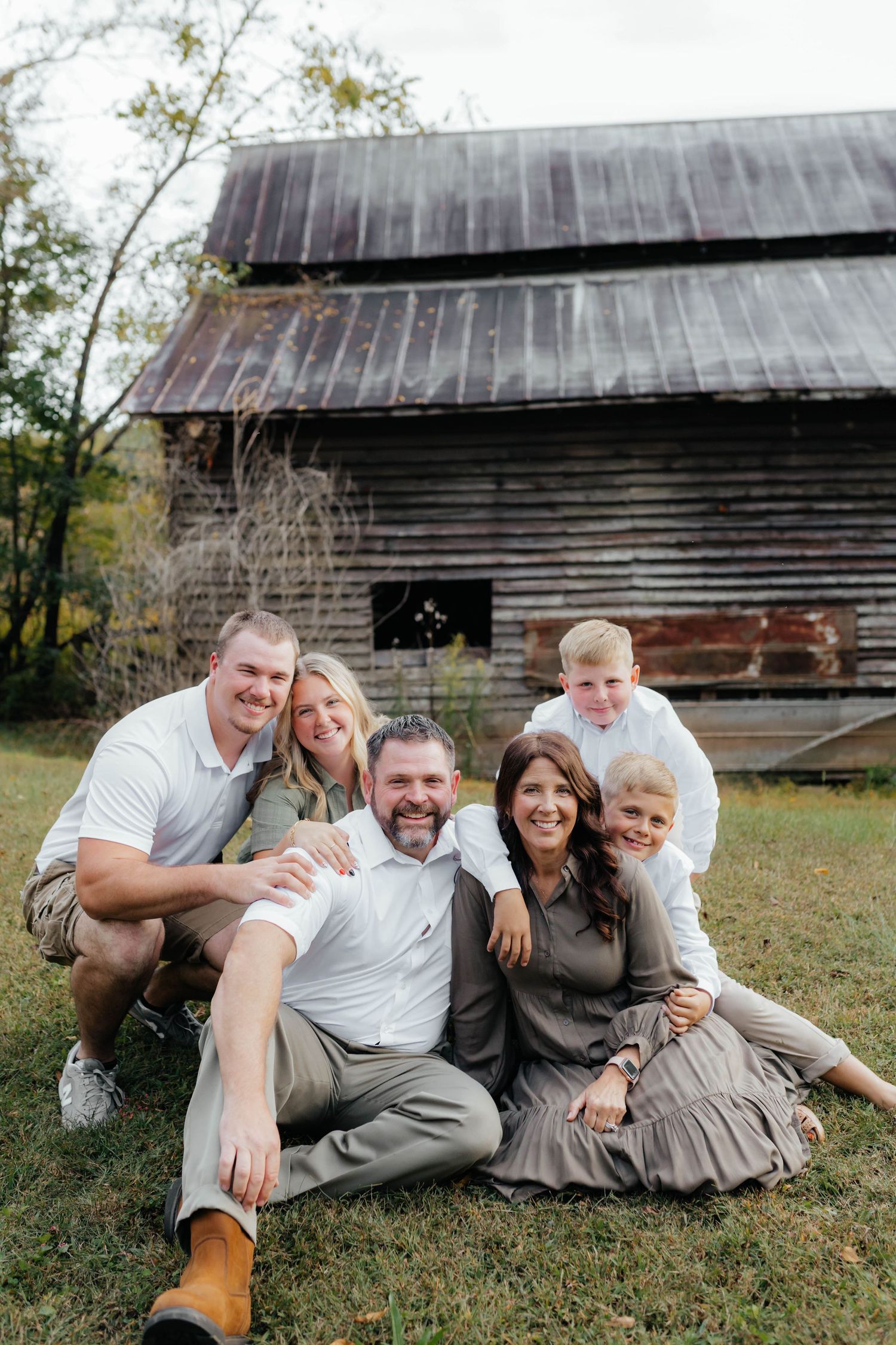 A family relaxes on grass for casual portraits in front of a weathered wooden barn.
