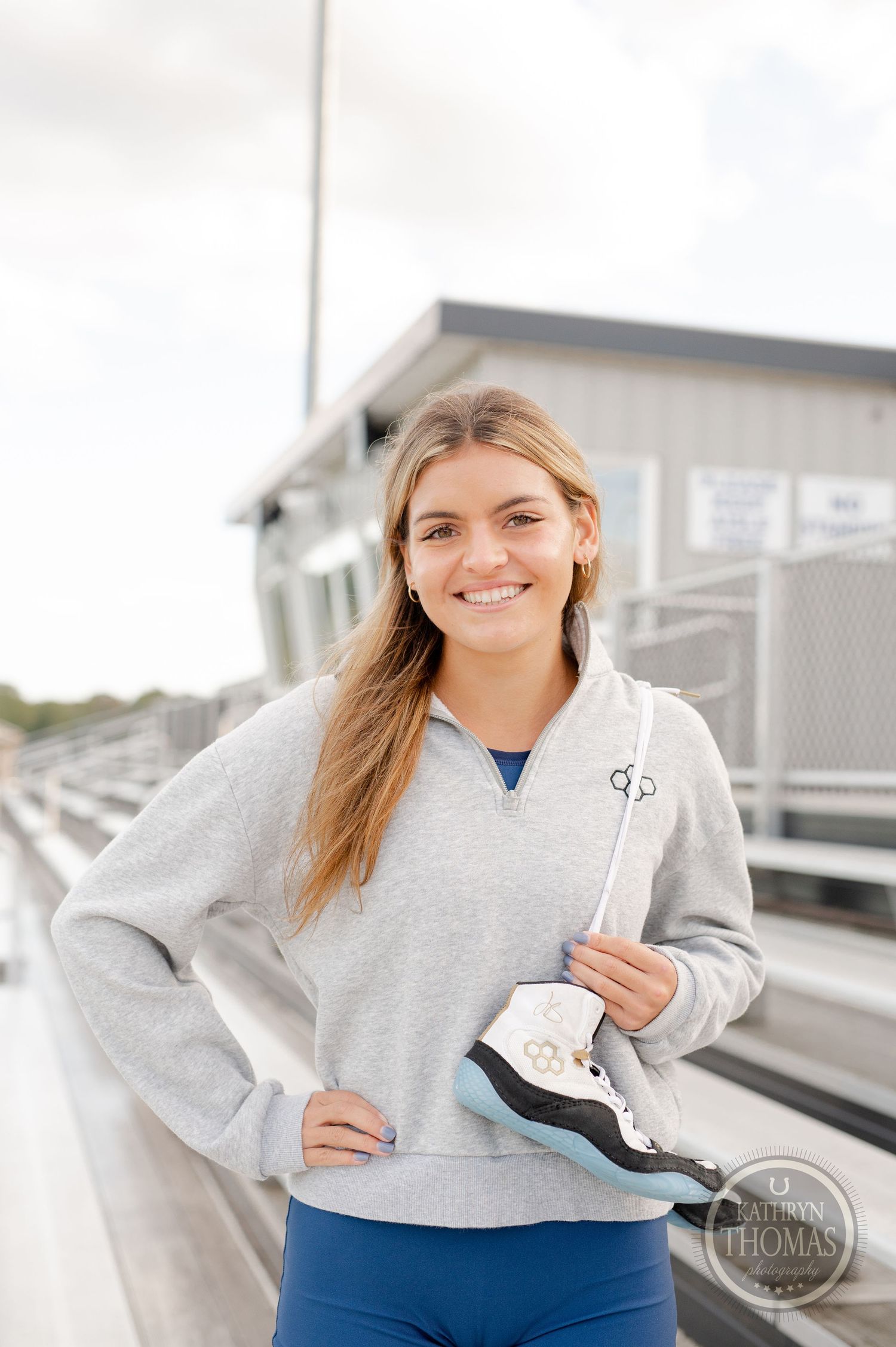 Athlete in grey quarter-zip sweatshirt holds ice skates while standing on metal stadium bleachers.