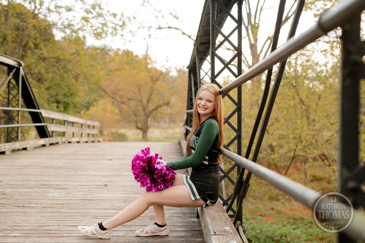 Cheerleader poses with pink pom poms while sitting on wooden bridge railing.