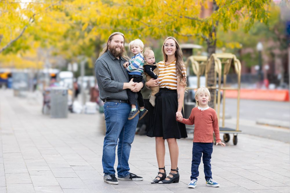 Family standing together on city sidewalk during autumn with yellow trees in background.