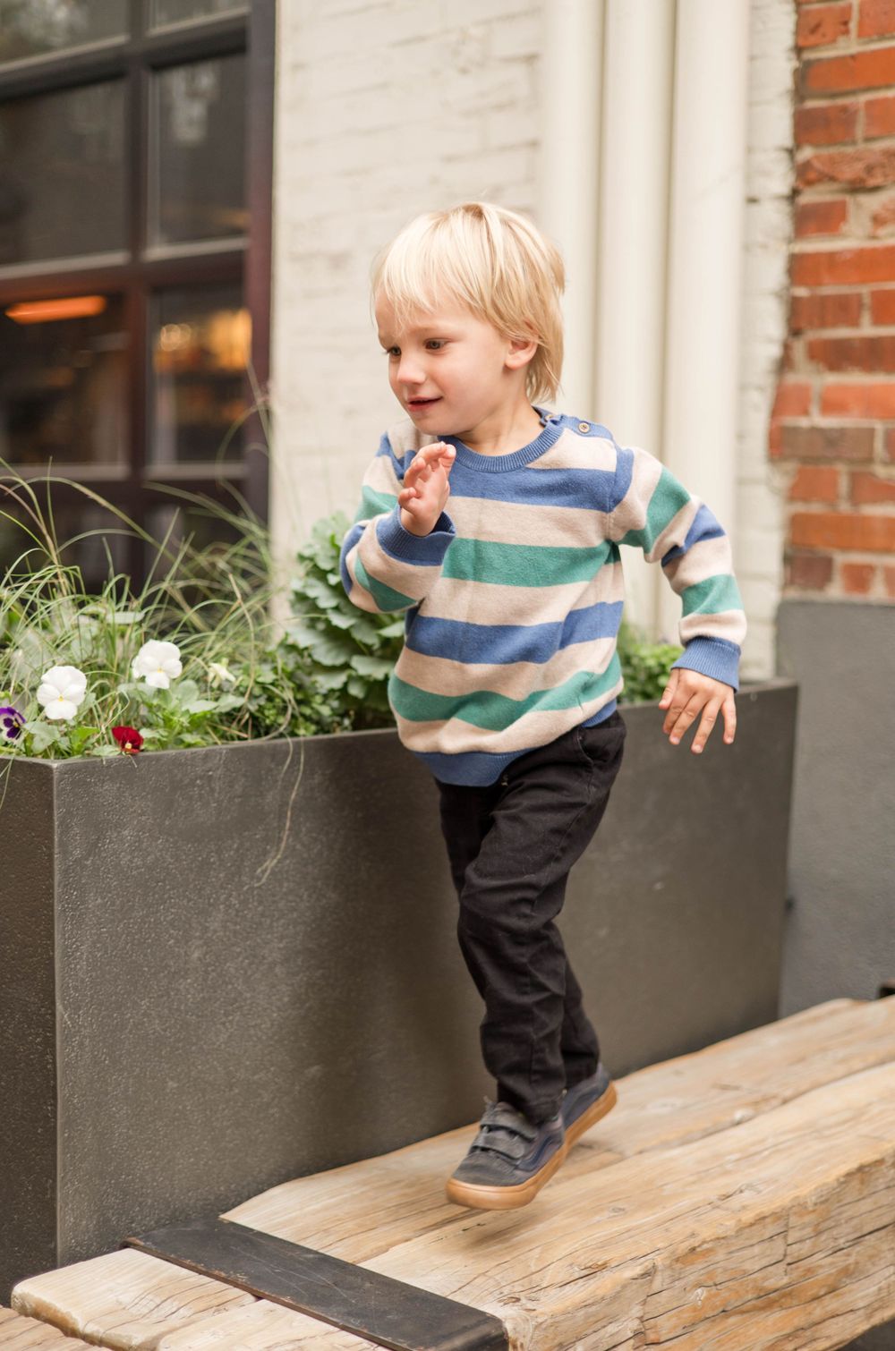 A child in a striped sweater plays on a wooden platform near planters.