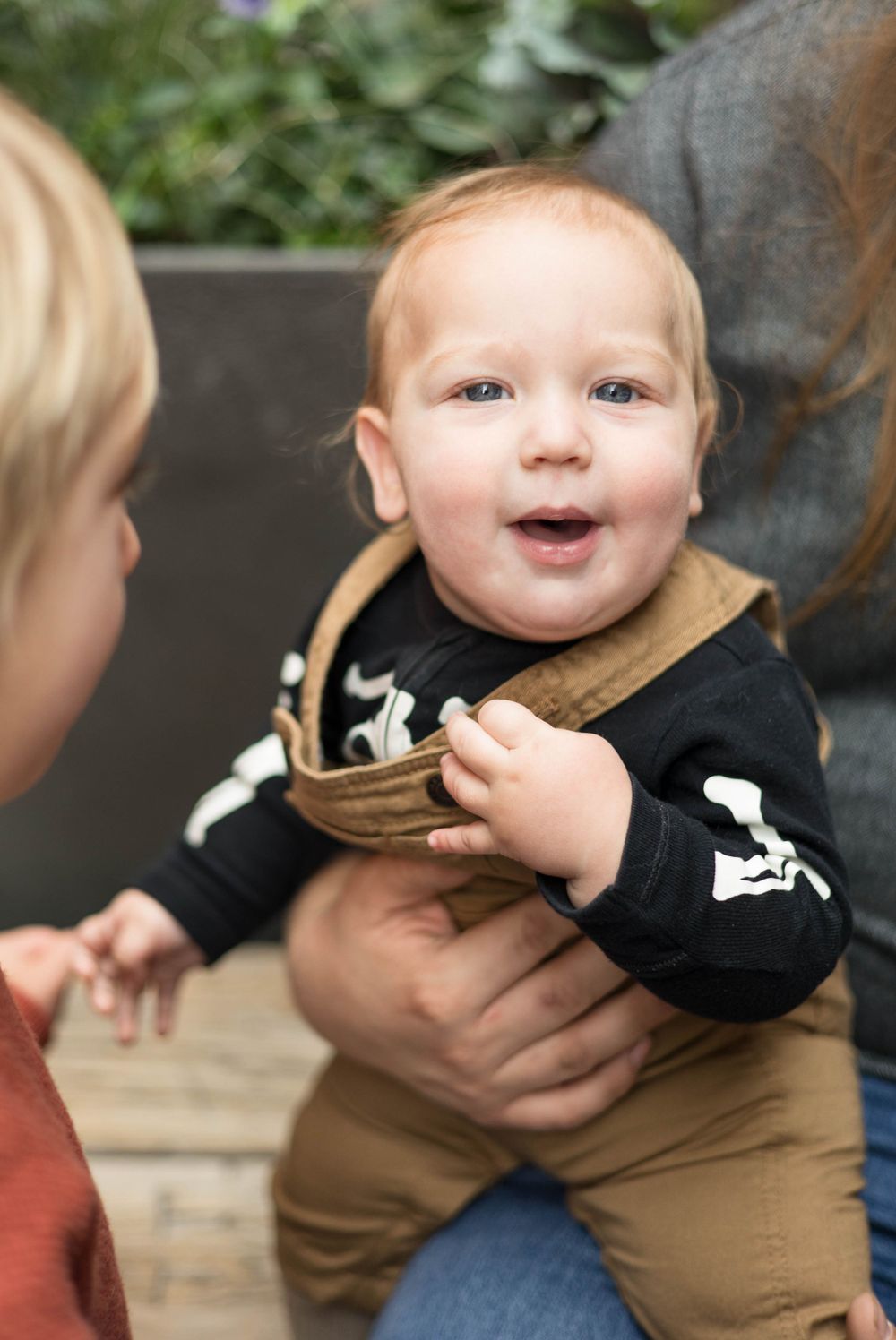 A happy baby in overalls and black shirt interacts with others outdoors.