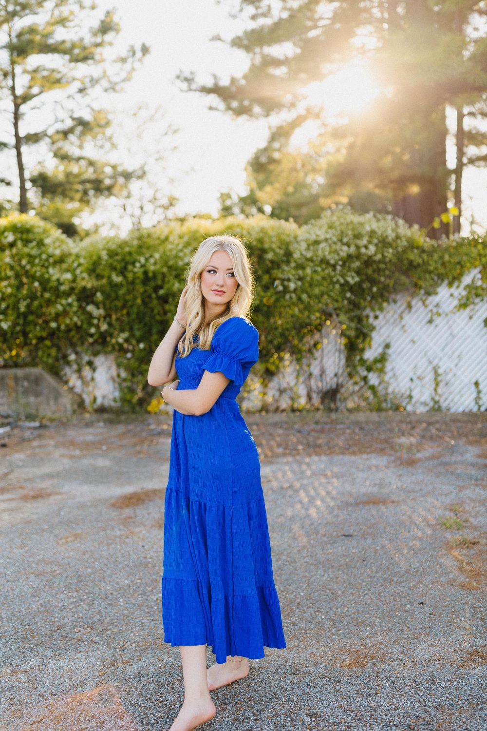 Person in a vibrant royal blue maxi dress poses outdoors at sunset with trees and a white fence in the background.