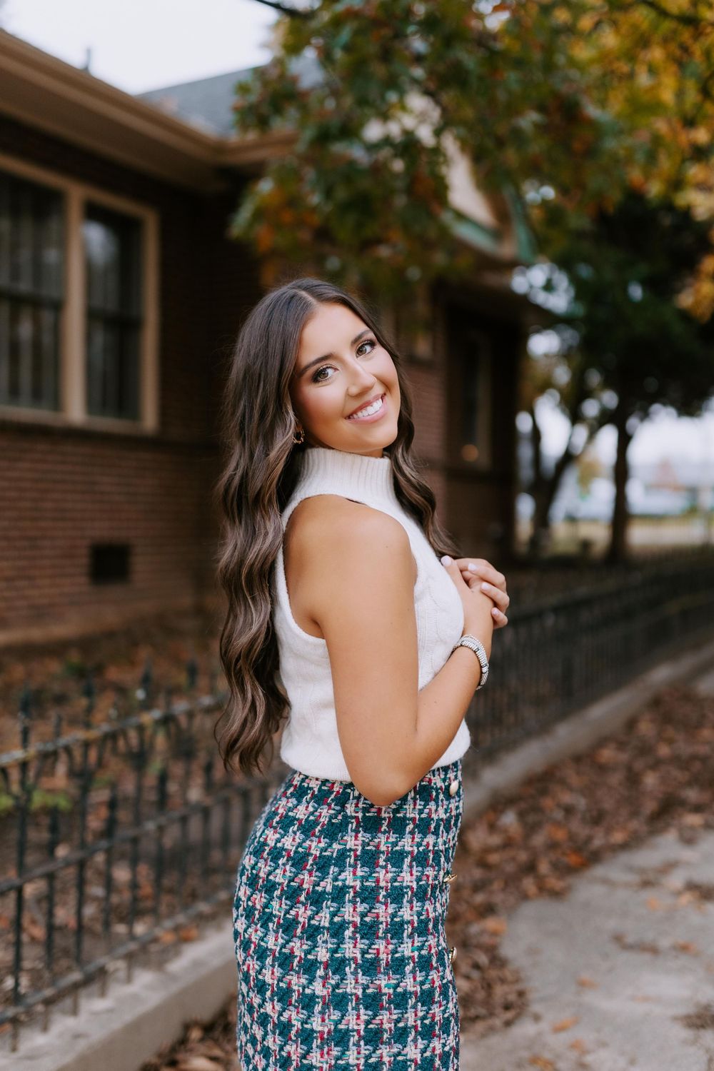 Woman in white sleeveless top and patterned skirt poses outdoors in autumn with brick building and foliage background.