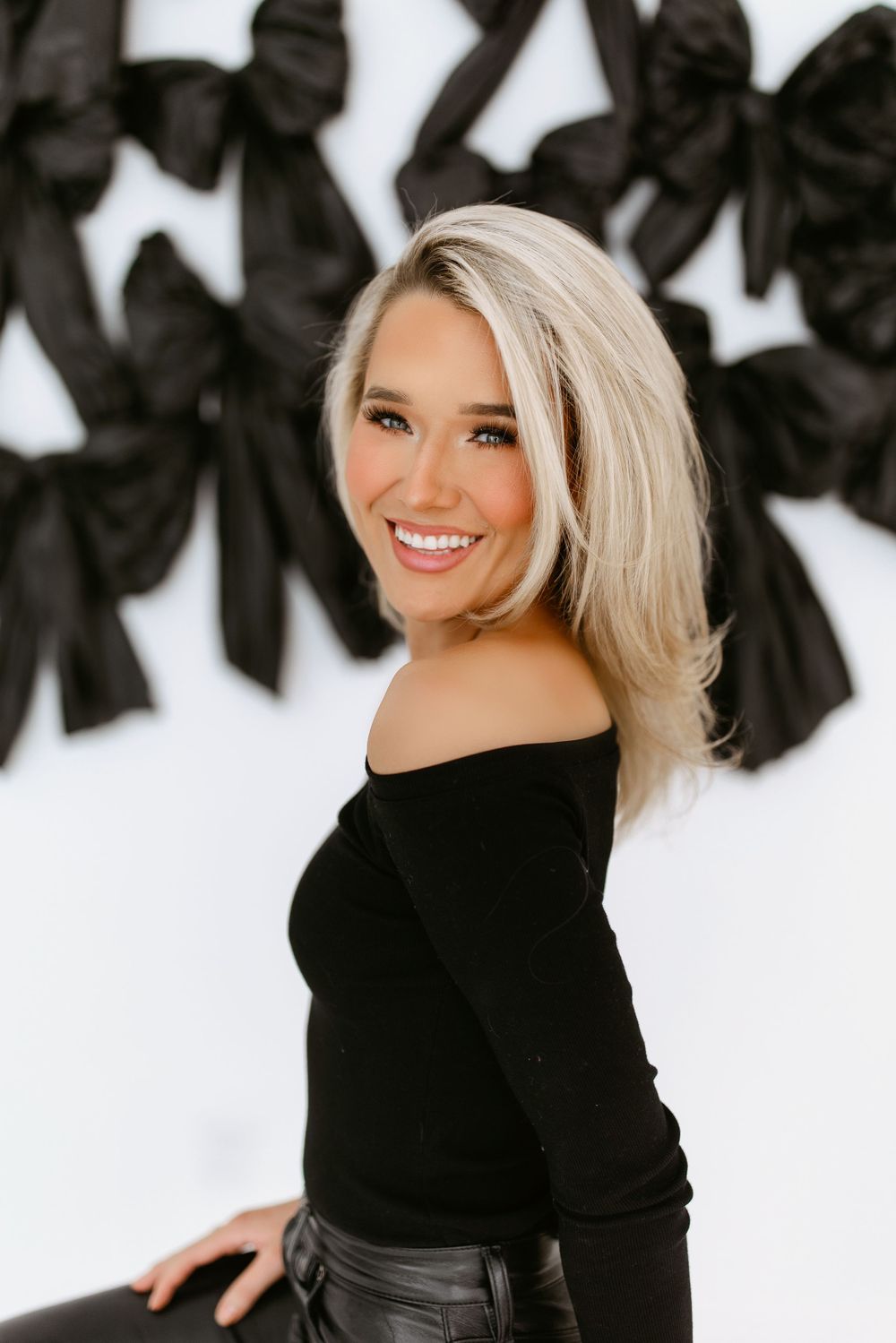 Portrait against white wall with black bow decorations featuring off-shoulder black sweater and platinum blonde hair styling.