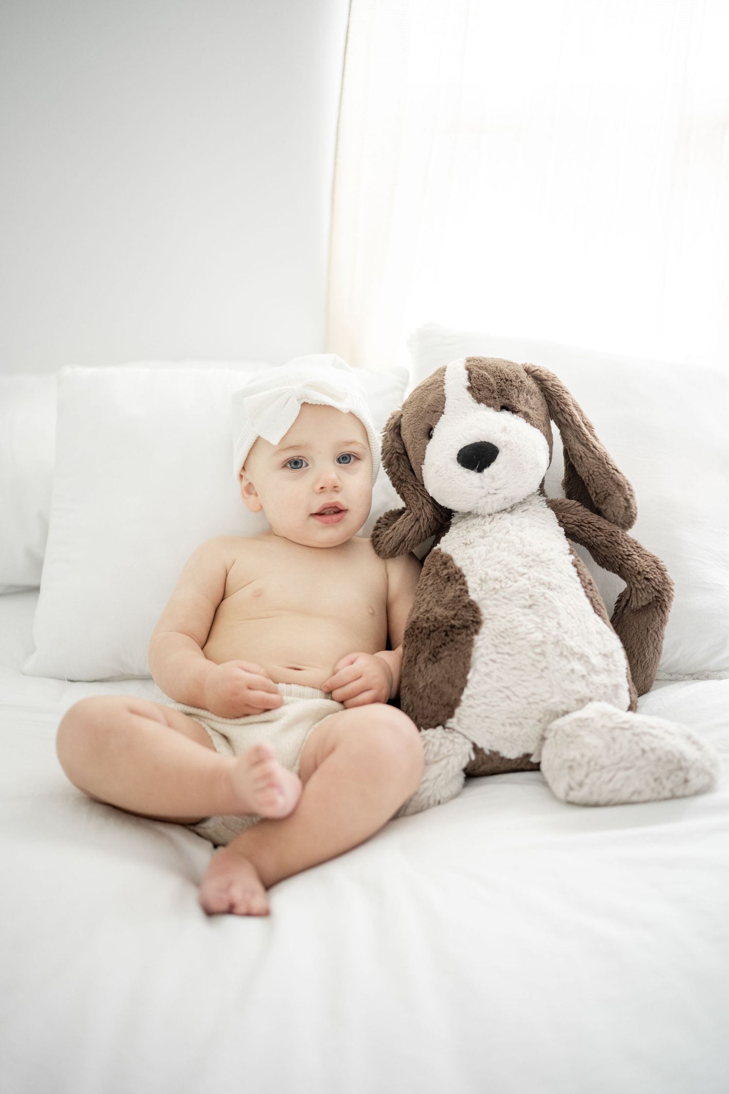little girl posed on white bed with favorite stuffed animal dog beside her.  girl is in bloomers with white hat