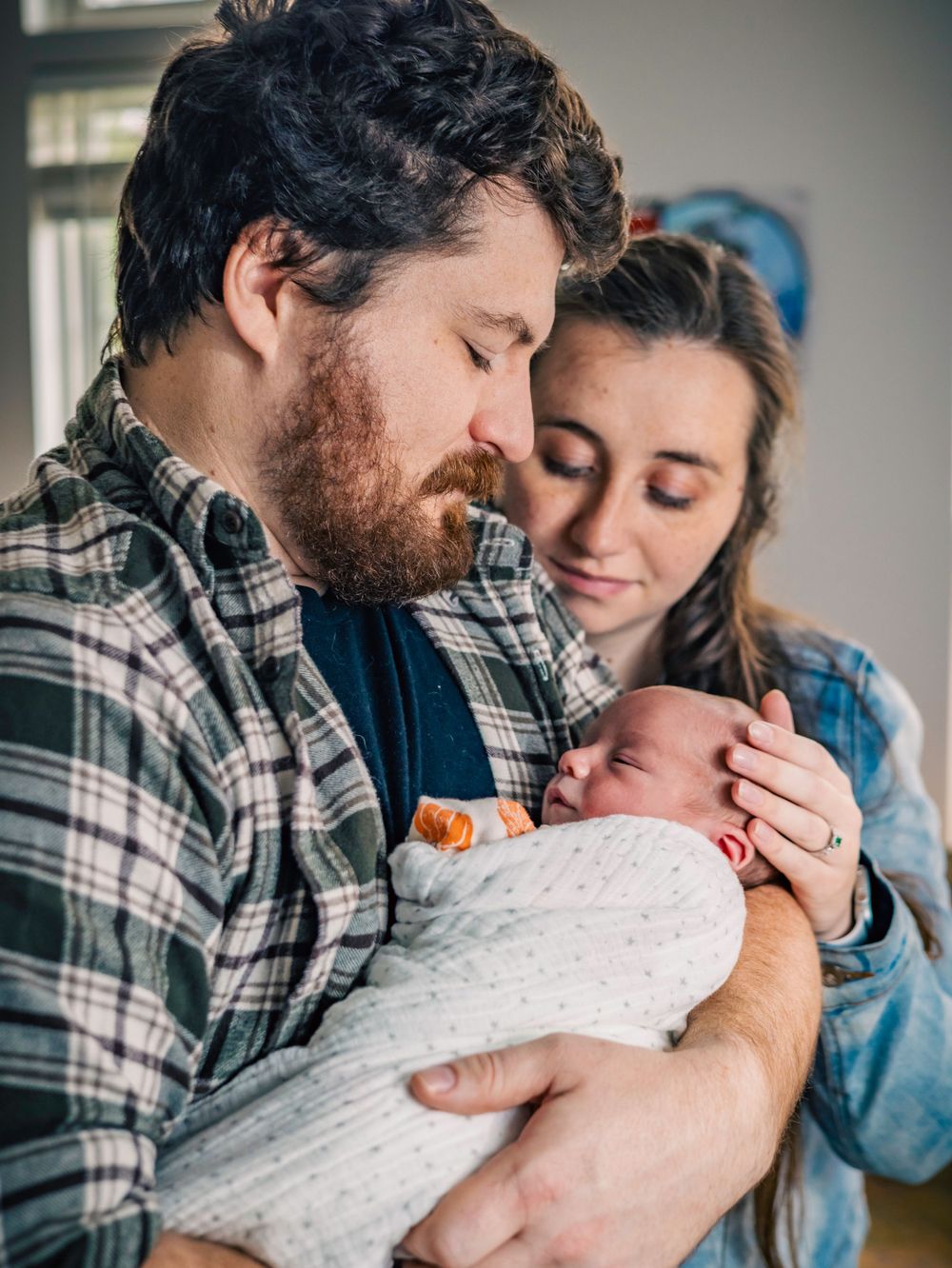 New parents in plaid and denim sharing intimate moment with swaddled newborn.