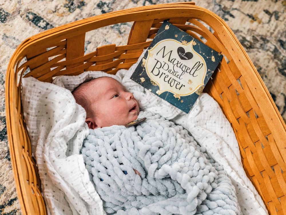 Sleeping newborn in white wrap rests in woven basket with milestone card.