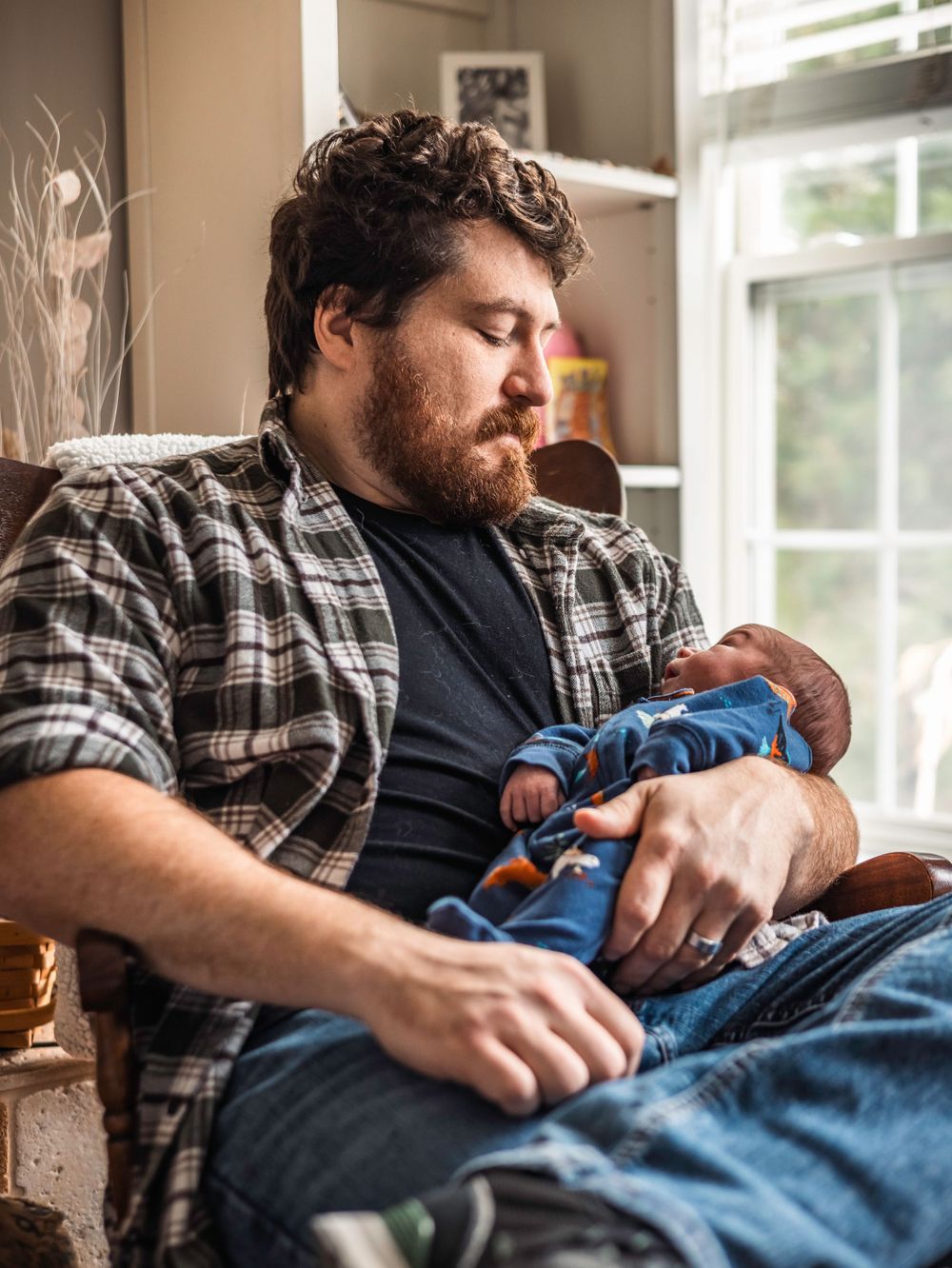 Parent in plaid shirt holds sleeping baby while sitting by window.
