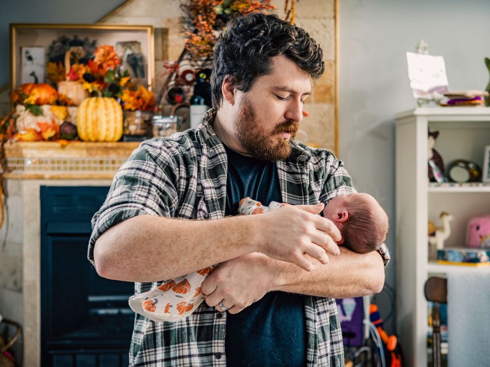 Parent in plaid shirt holds swaddled newborn in autumn-decorated living room.