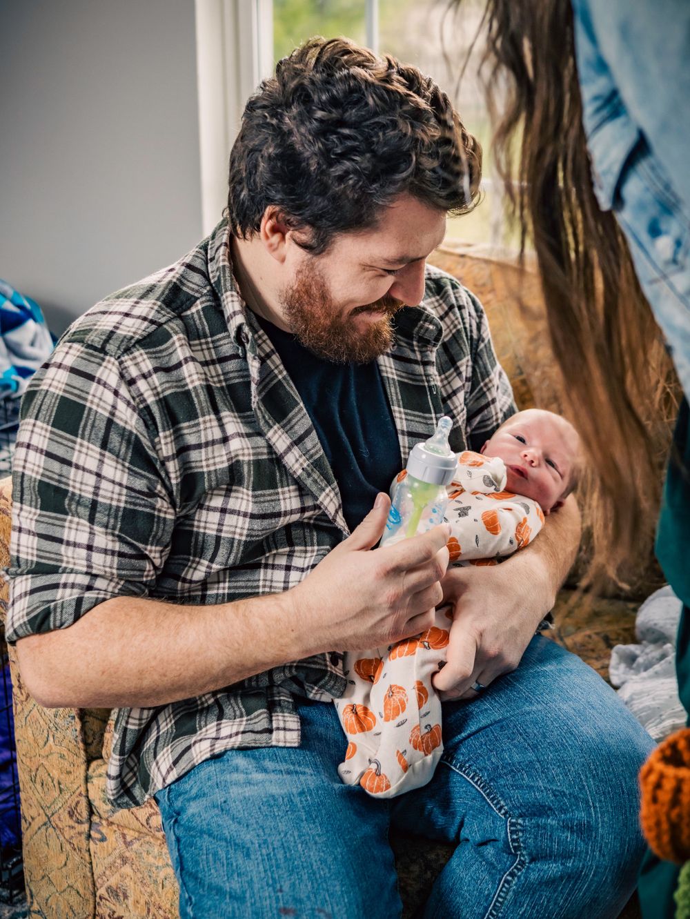 Adult in plaid shirt tenderly feeds newborn swaddled in patterned blanket.