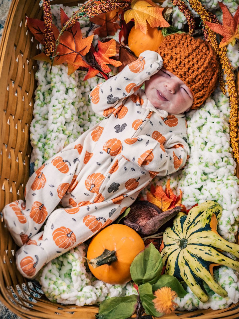 Infant in pumpkin-print onesie and orange hat lies in basket with fall decorations.