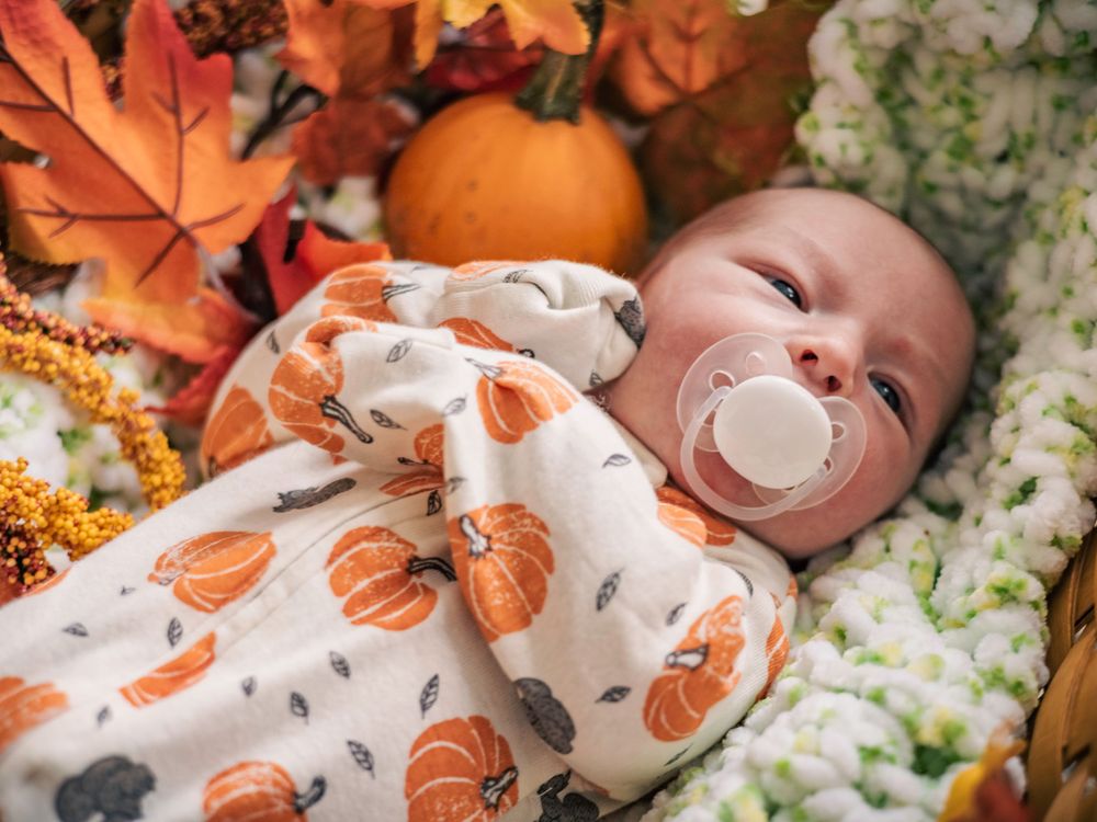 Sleeping infant in pumpkin-print outfit surrounded by autumn decorations in wicker basket.
