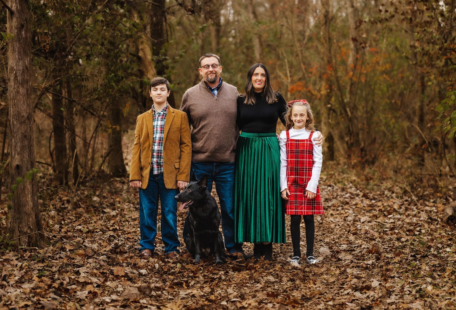 Family of four with black dog posing for formal portrait in autumn woods.