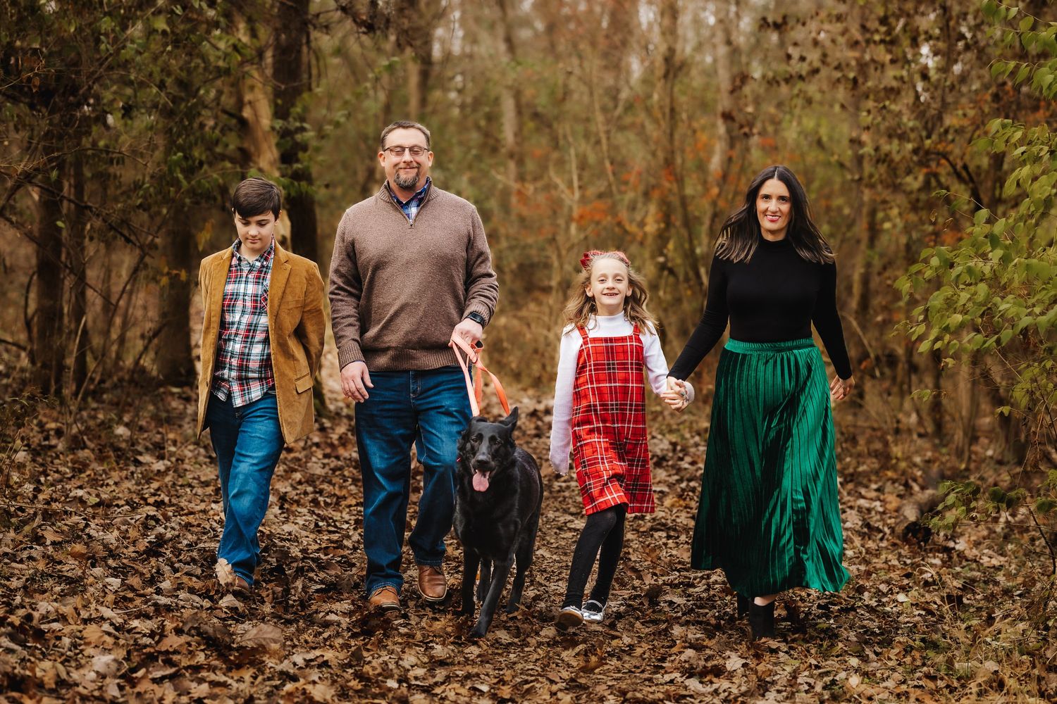 Family walking through autumn forest with black dog on fallen leaves.