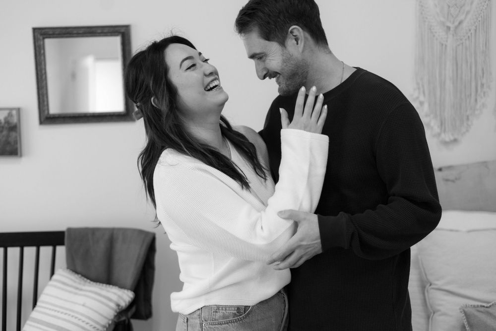A couple shares a candid moment of laughter together in a living room, captured in black and white photography.