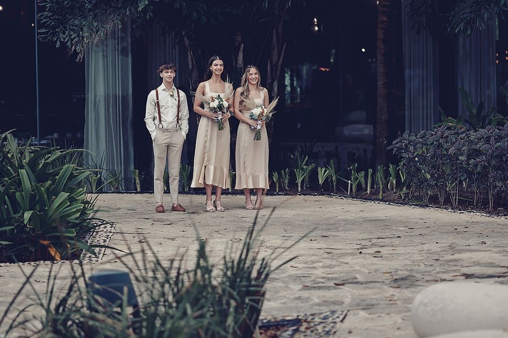 Wedding party members in champagne colored attire stand together at a garden venue.
