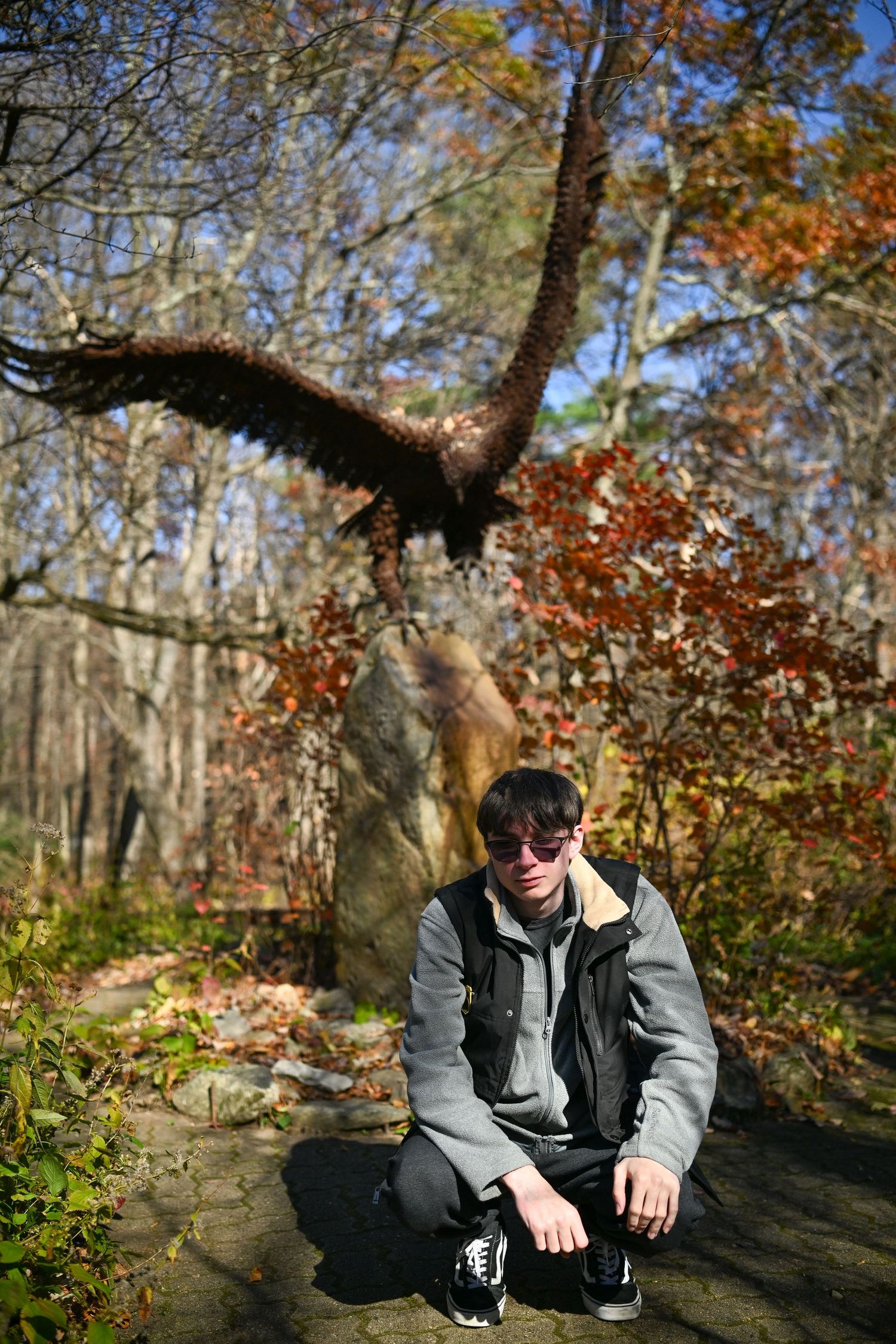 Photograph of a young man looking serious and kneeling in front of a large metal sculpture of a bird in flight