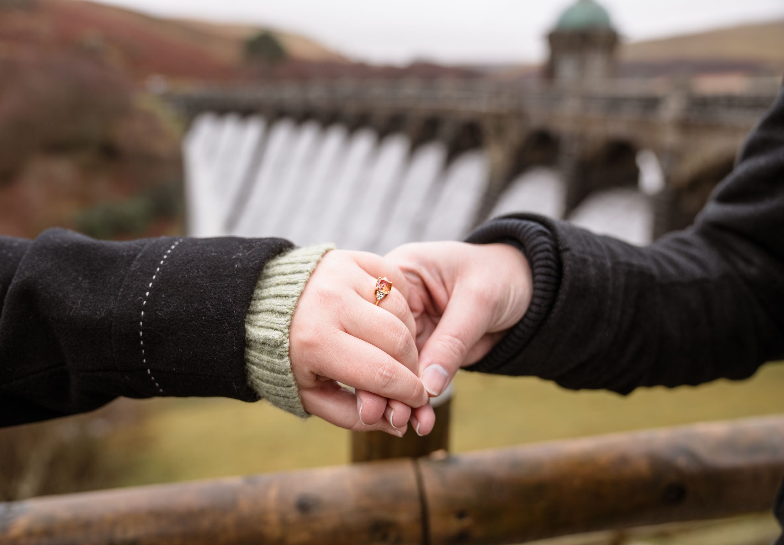 Two people holding hands together in front of a dam or reservoir with a blurred background.