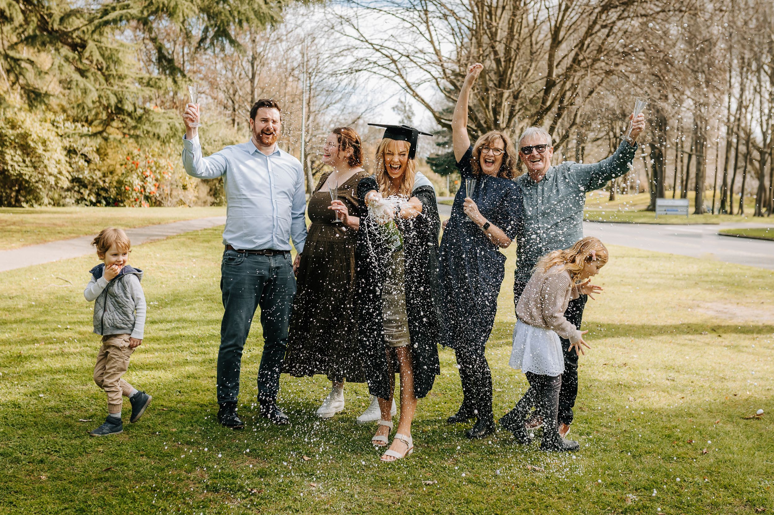 Woman in graduation cap and gown with her family at the Canterbury University campus spraying a bottle of champagne on her graduation day.