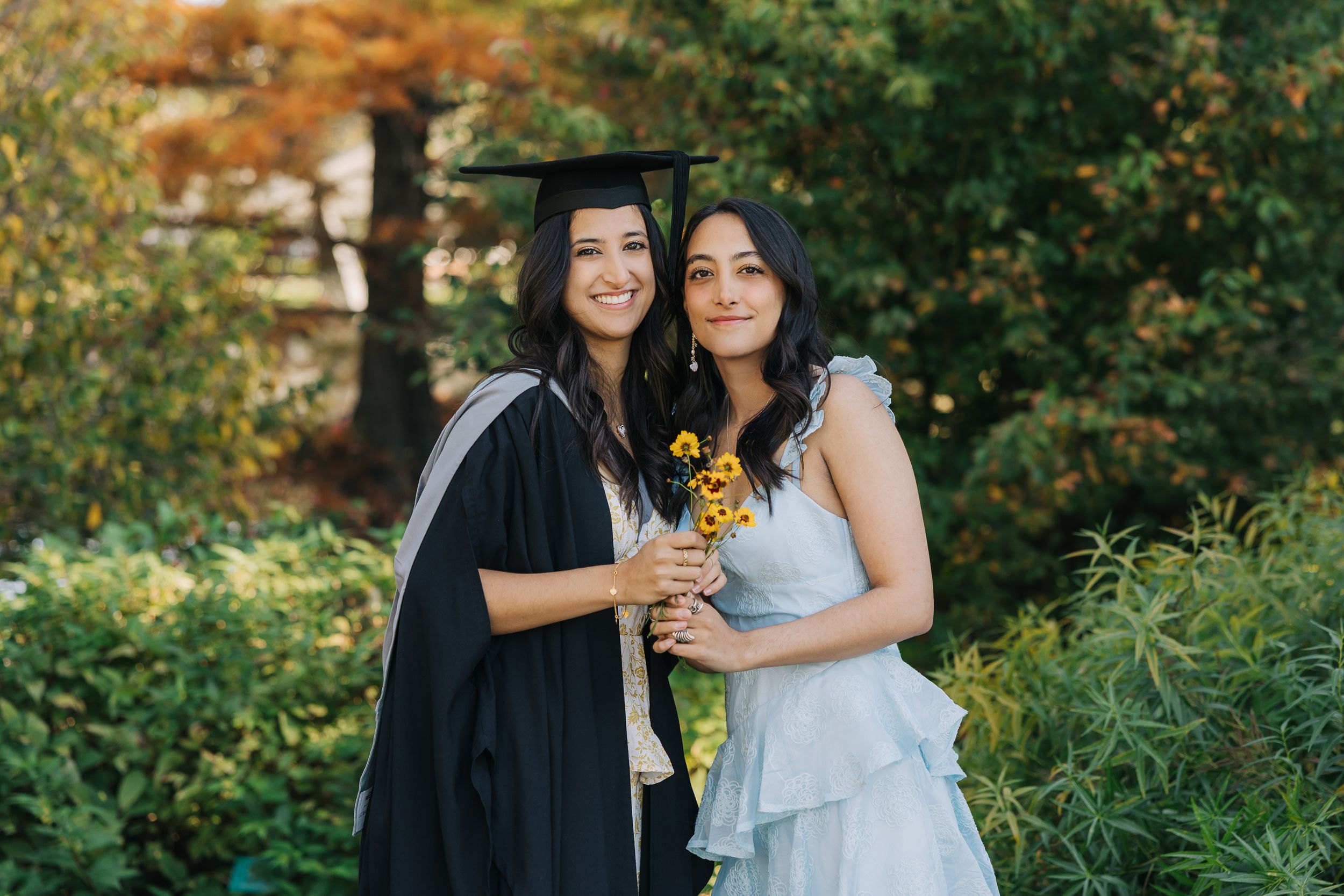 Female in her graduation cap and gown, cuddling with her sister in the Christchurch Botanic Gardens.