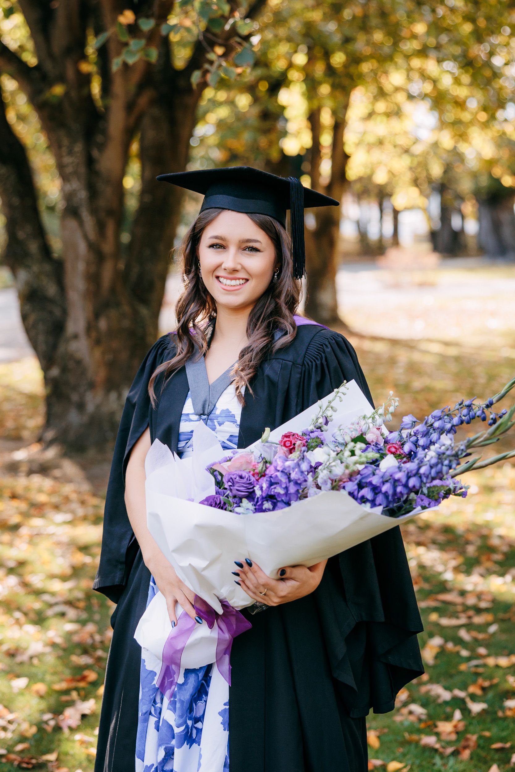 Young woman in graduation gown and cap, holding a bunch of flowers, trees in background, in the Canterbury University campus, Christchurch.