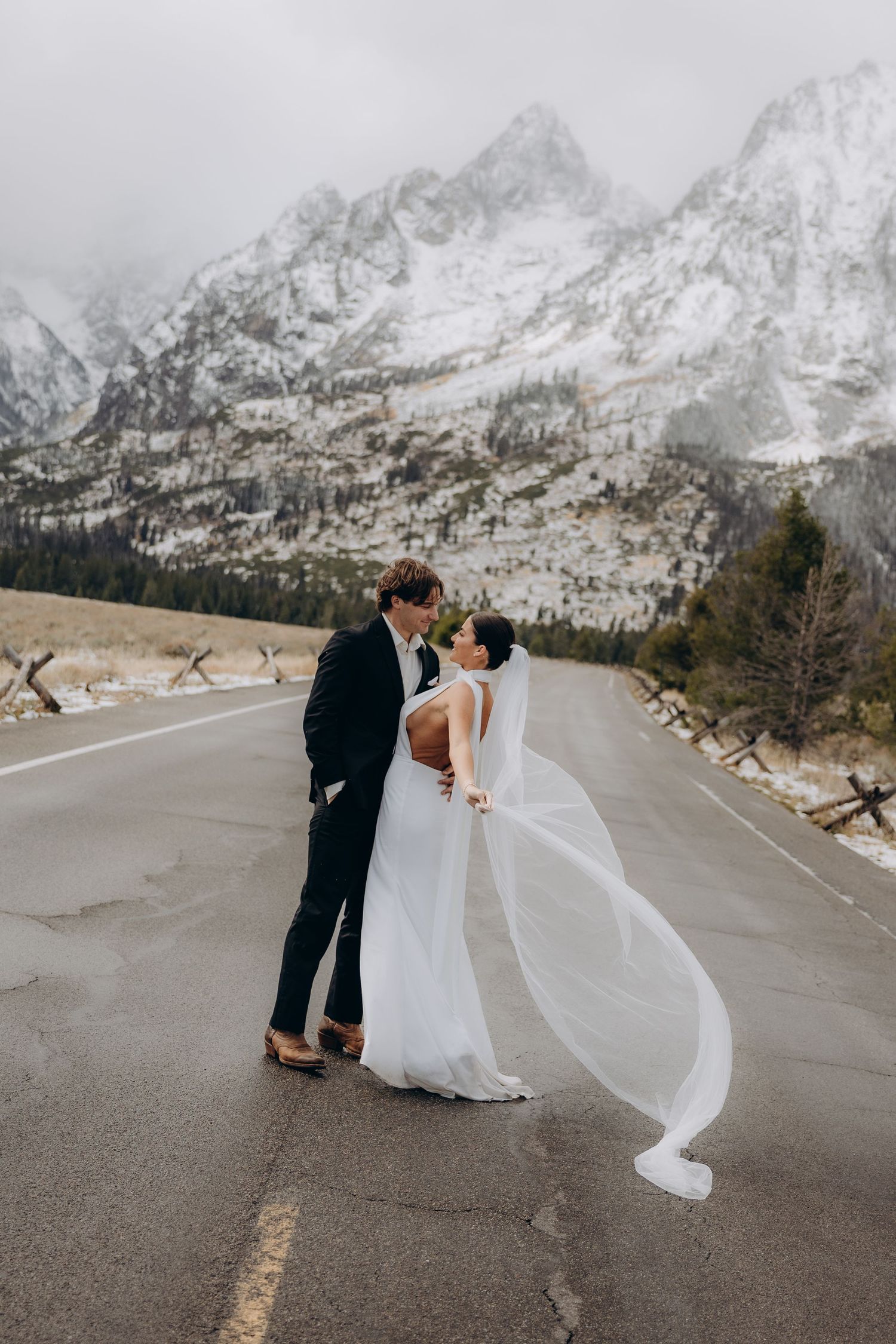 A couple in wedding attire stands on a mountain road with snow-capped peaks and dramatic cloudy skies in the background.