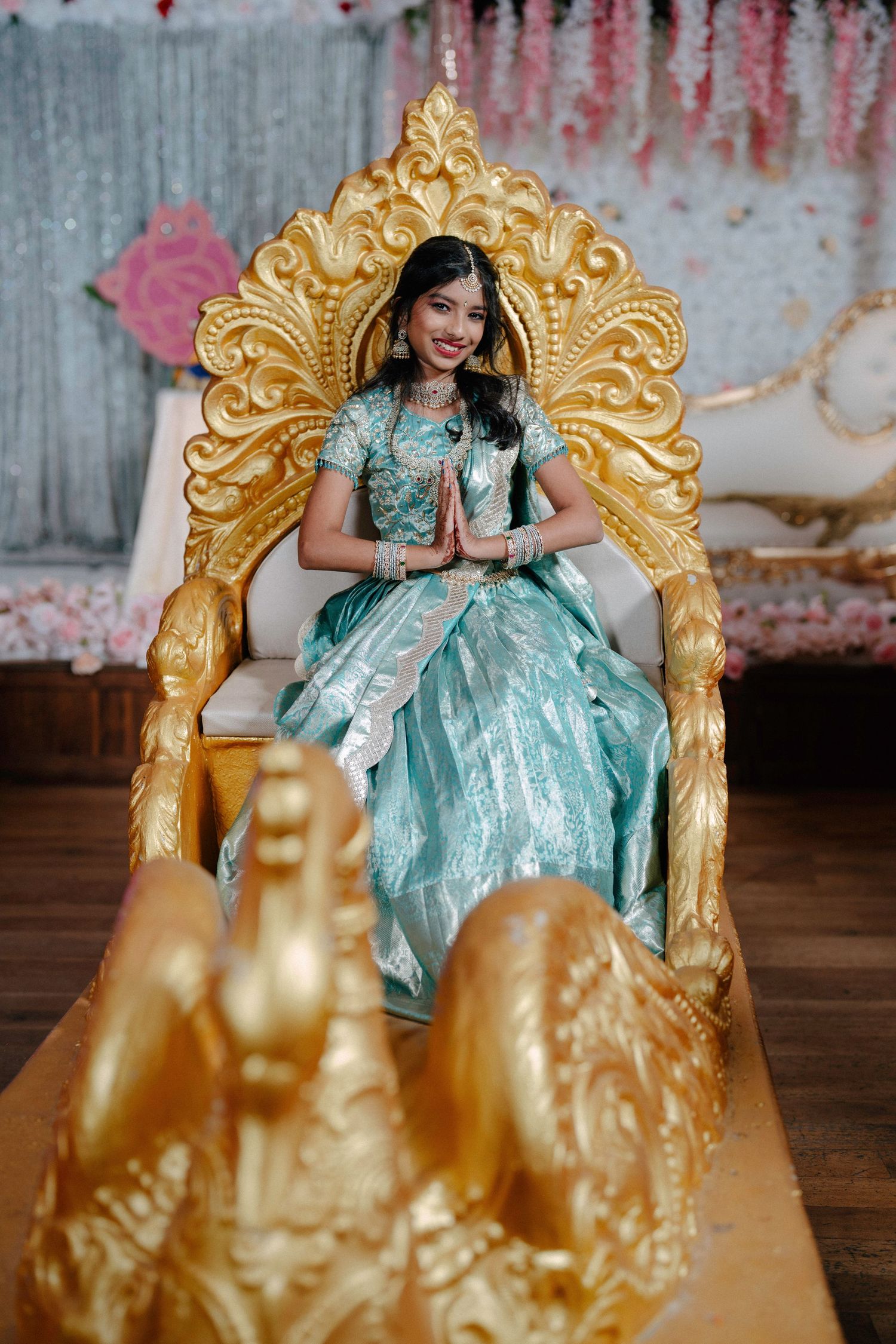 Elegant poses on an ornate gold throne with mint green attire against a pink floral backdrop.