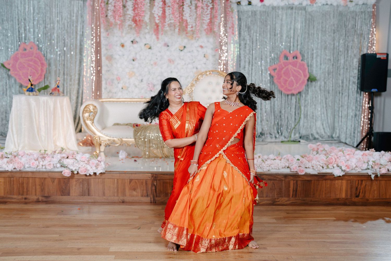 Two dancers in bright orange dresses perform a traditional dance routine at an indoor venue.