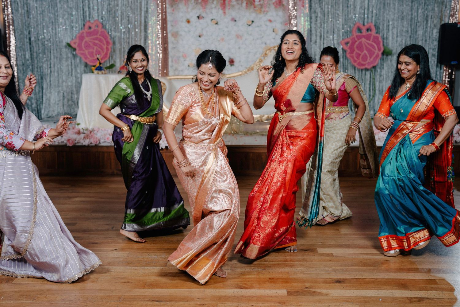 Group celebration with colorful traditional Indian saris and dresses on wooden floor.