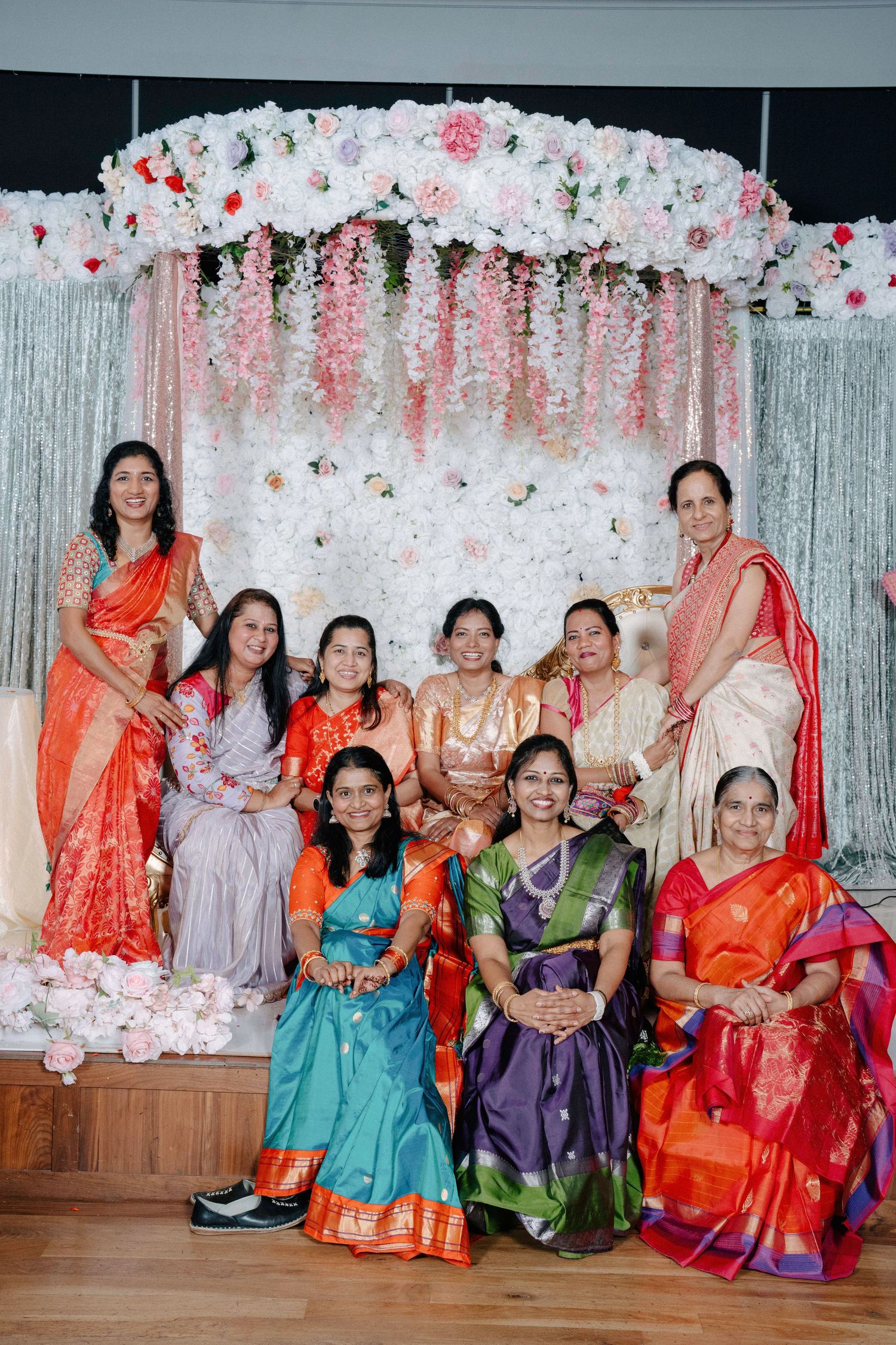 A group in colorful traditional Indian saris and outfits posing together in front of a floral wall decoration.