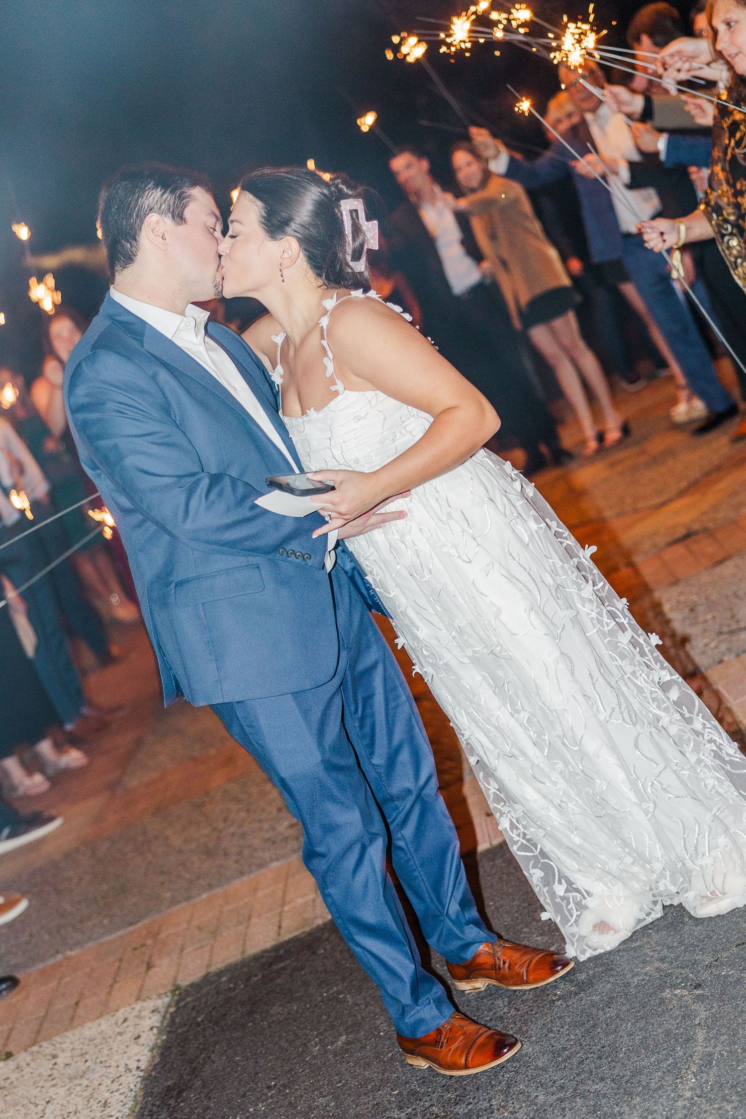 A romantic sparkler exit with a couple sharing a kiss at their wedding celebration while guests hold sparklers in the background.
