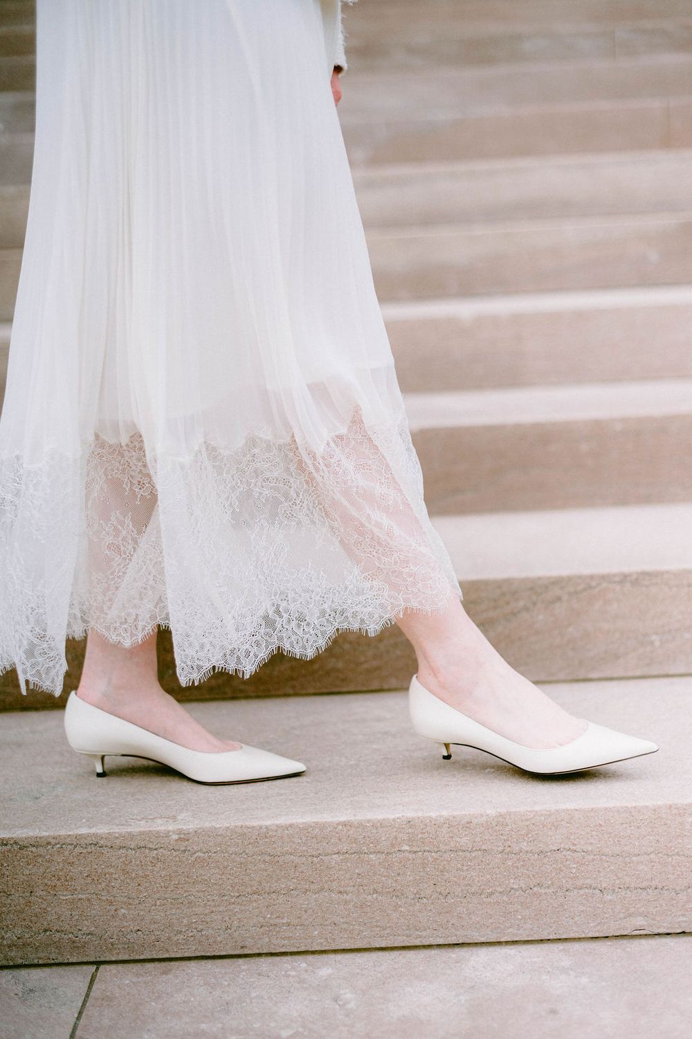 Close-up view of elegant white bridal shoes peeking beneath a delicate lace wedding dress hem.