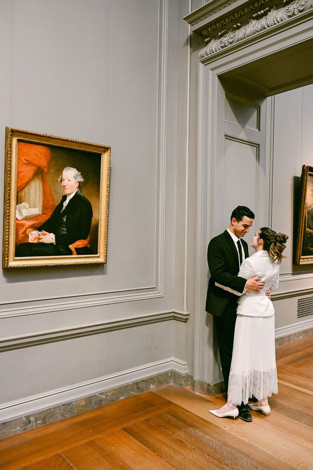 A couple shares a romantic moment in an elegant museum gallery with portrait paintings on the wall.