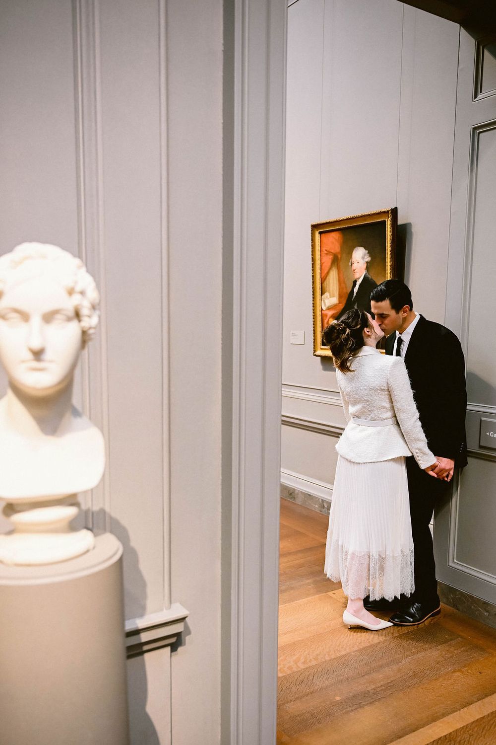 A couple embraces in front of white gallery walls with artwork and a sculptural bust visible.
