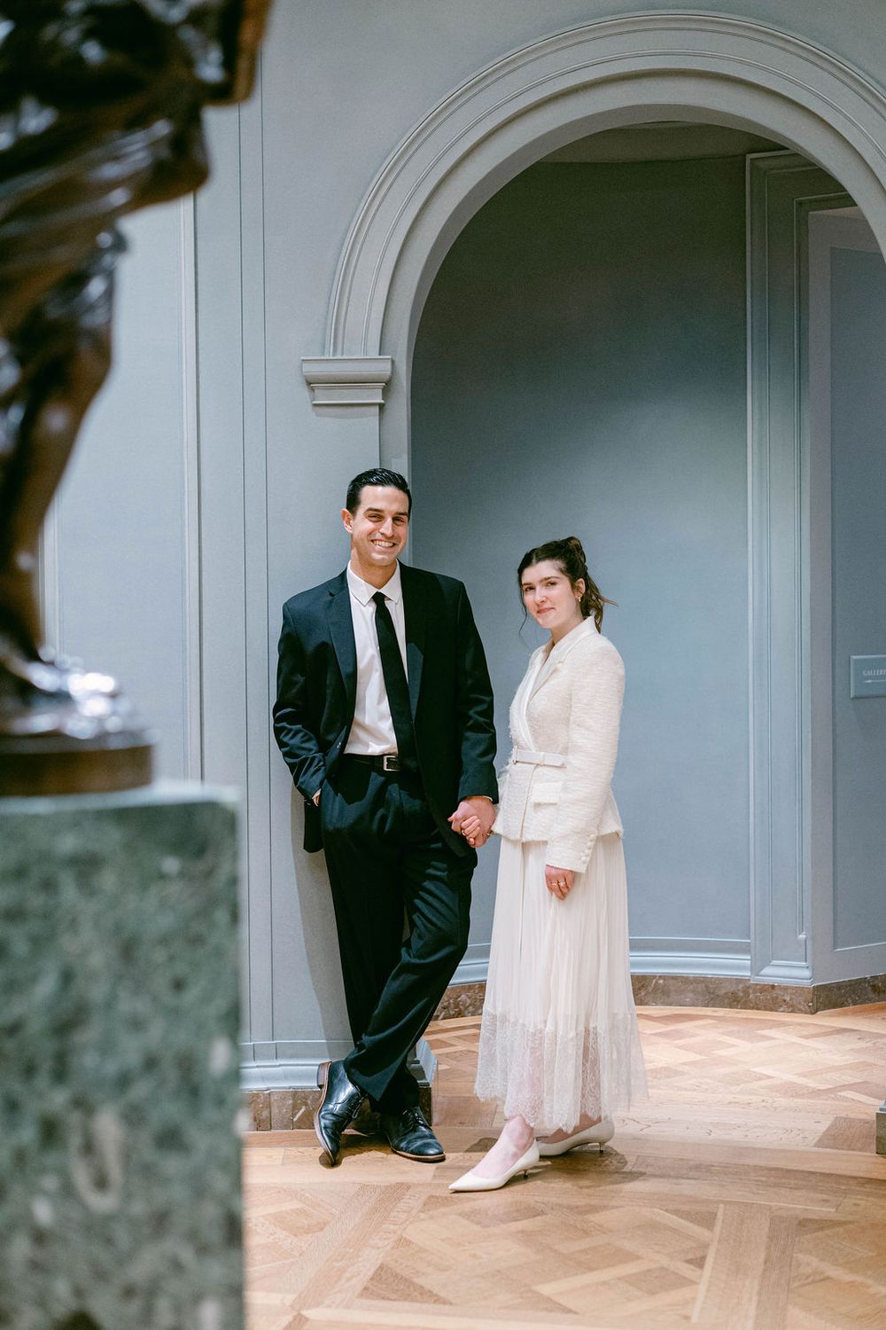 A couple in formal attire poses in an elegant archway with classic architectural details and parquet flooring.