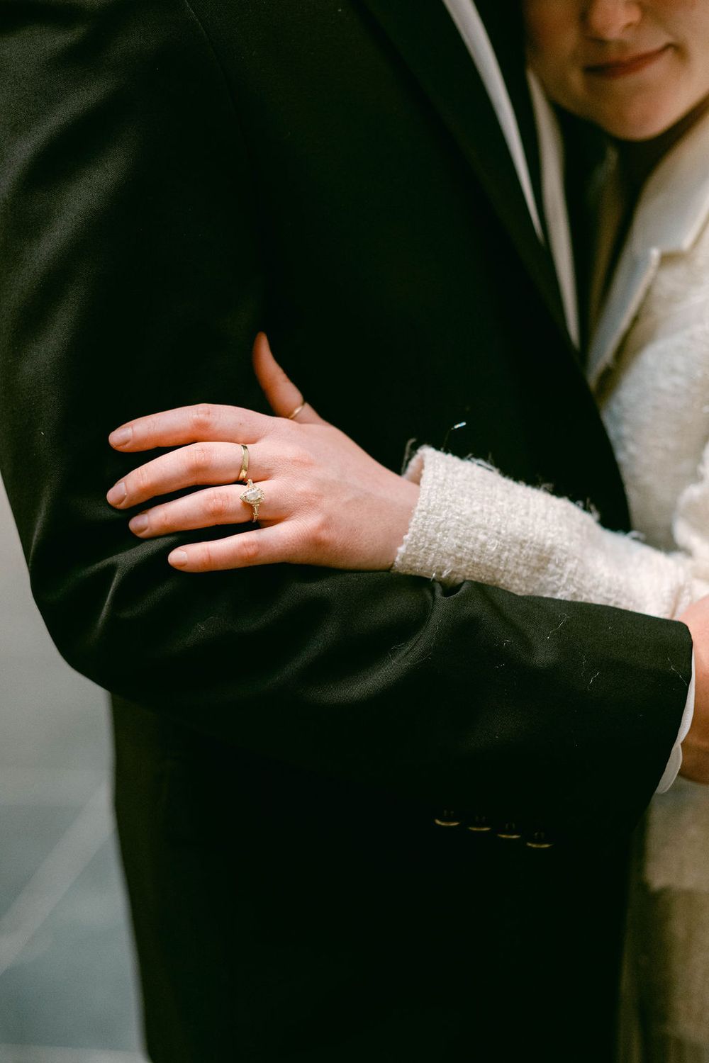 Close-up of a bride's hand with wedding ring resting on groom's black suit jacket sleeve.