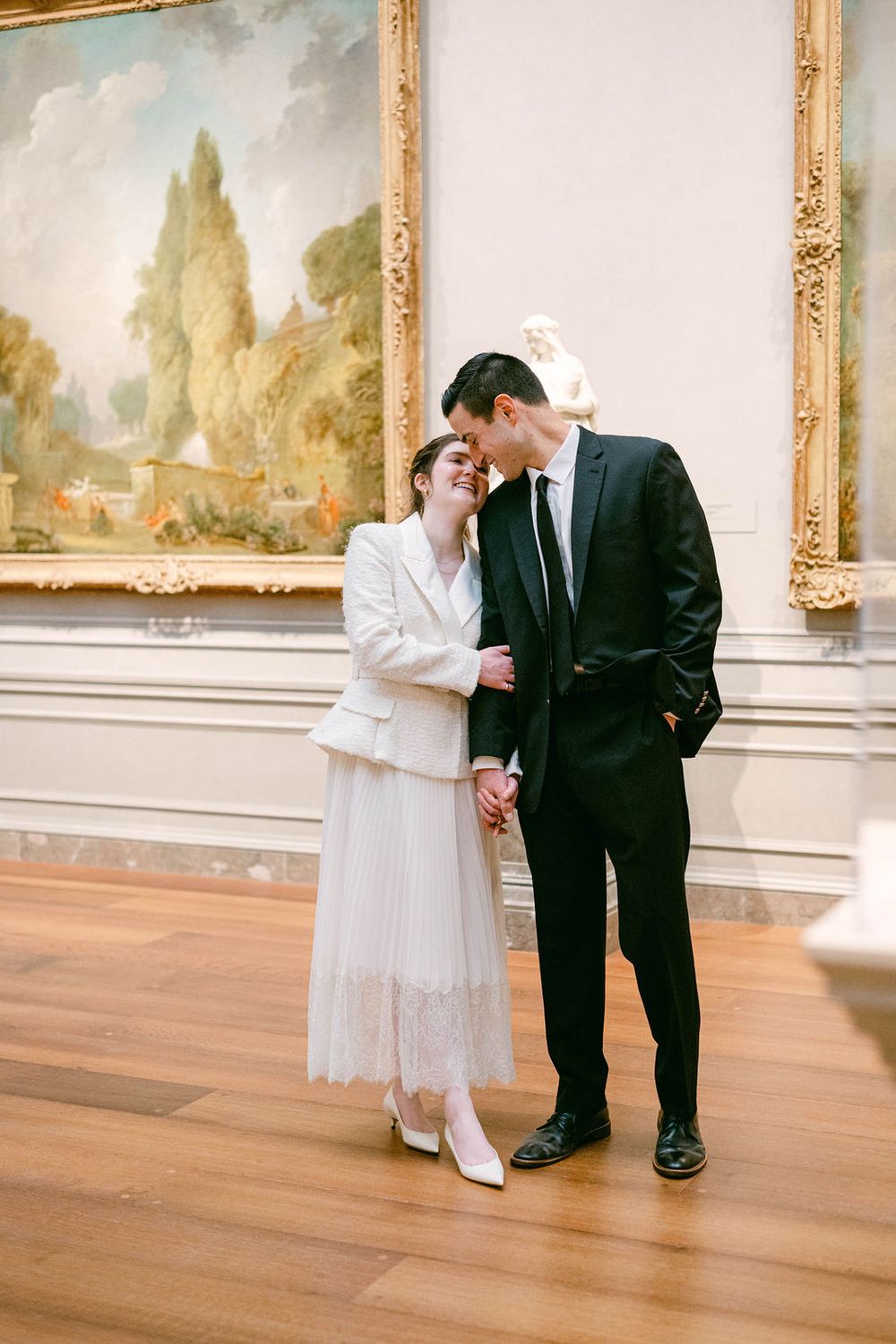 A couple in formal attire shares an intimate moment while viewing artwork in an elegant gallery with gold-framed paintings.